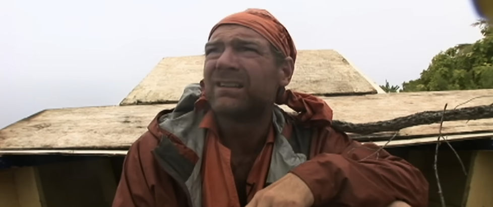 a screenshot from the Cook Islands episode of Survivorman with Les sitting in an abandoned boat facing the camera and looking annoyed by the rain