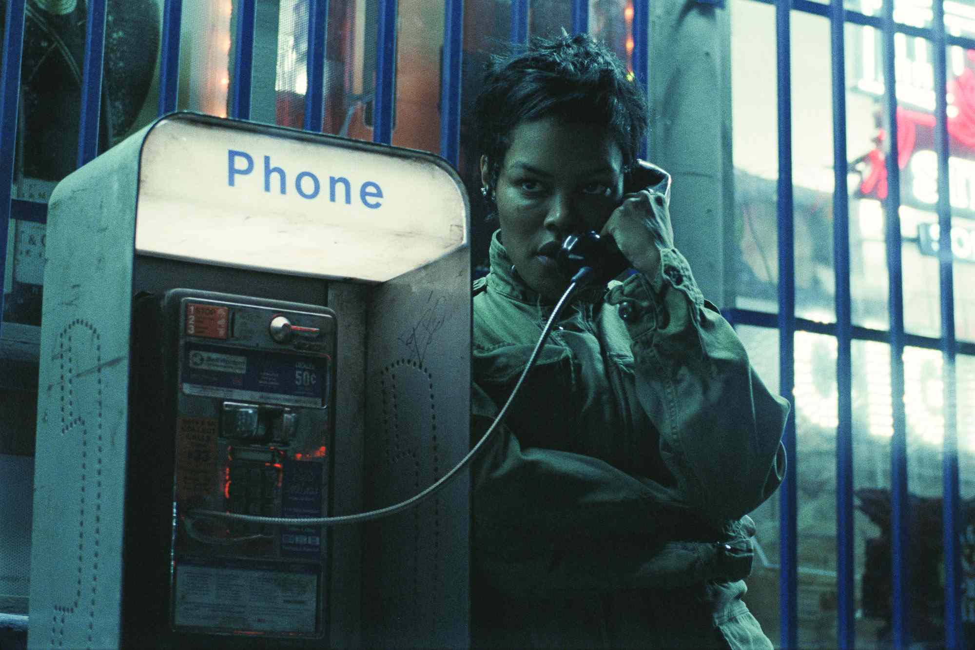 A still from One Battle After Another shows Teyana Taylor as Perfida Beverly Hills surreptitiously using a payphone while scanning the area around her, eyes alert.