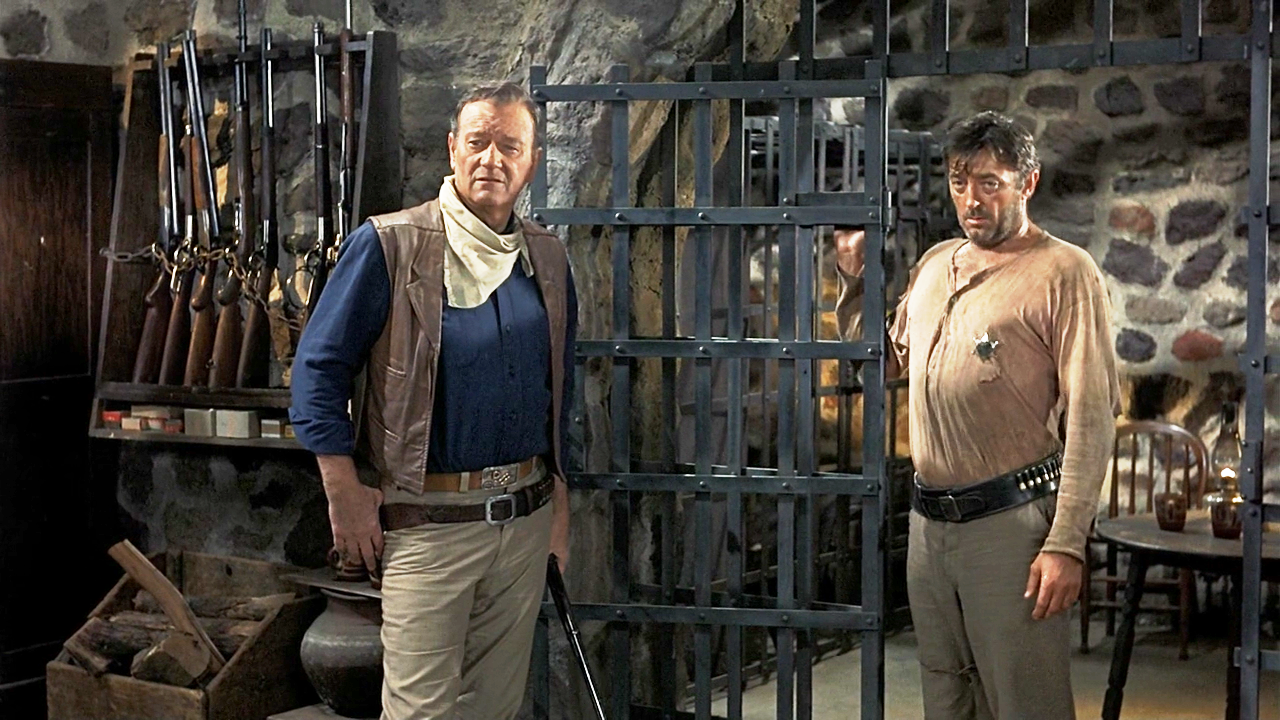 John Wayne and Robert Mitchum stand together in a rustic jail cell in this still from El Dorado, both looking more than a little worse for the wear.
