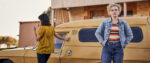 A still from the movie The Royal Hotel shows main characters Hanna and Liv standing in front of a retro car in a parking lot. Hanna stares somewhere past the viewer while Liv is turned away, gazing in another direction.