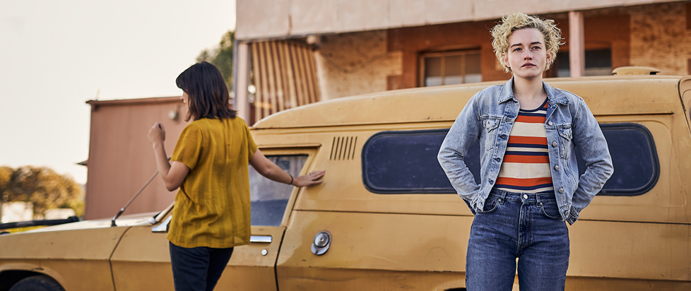 A still from the movie The Royal Hotel shows main characters Hanna and Liv standing in front of a retro car in a parking lot. Hanna stares somewhere past the viewer while Liv is turned away, gazing in another direction.