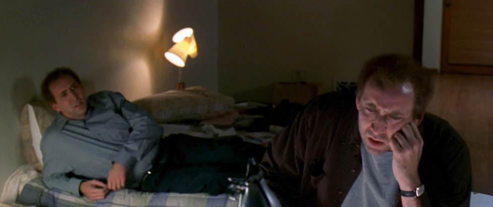 A still from Adaptation features Nicholas Cage in a dual role as twin brothers, mid conversation in a dimly lit living room.