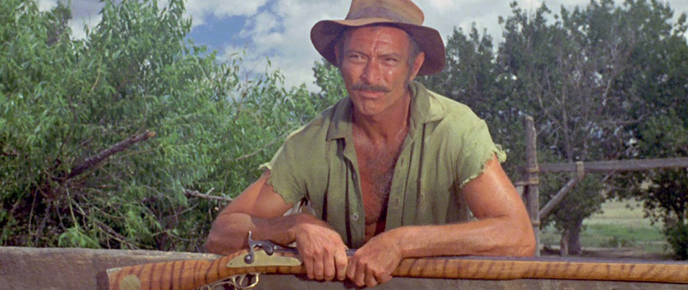 A still from Barquero of a rugged man leaning forward in the bright sun, a beat up hat on his head and a well-worn shirt worn open barely hanging on to his shoulders.