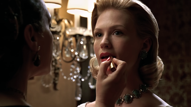 A screenshot of Mad Men with Better having her lipstick applied by another woman while standing in front of an elegant chandelier 
