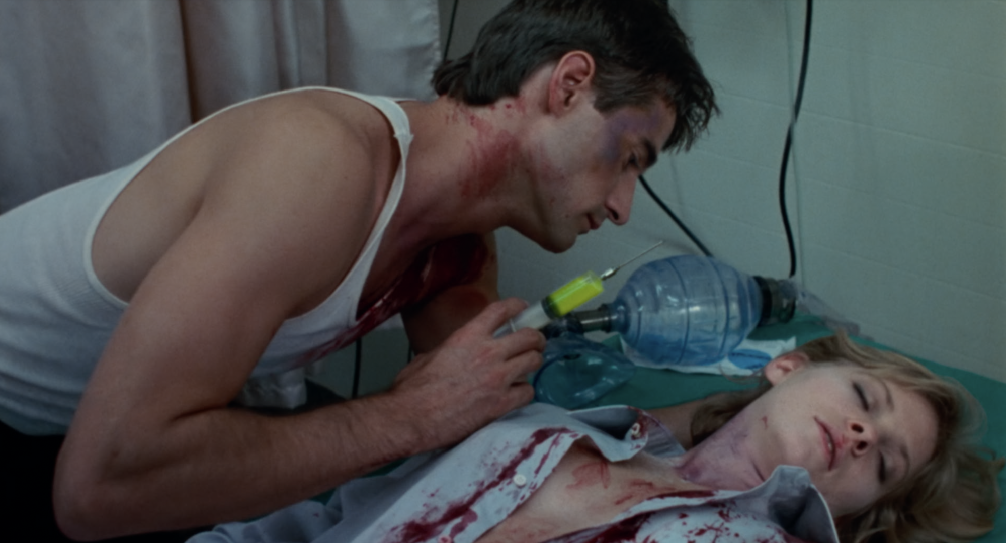 A bloodied man in an undershirt hovers menacingly over a prone woman covered in blood. He's holding a syringe full of bright yellow serum.