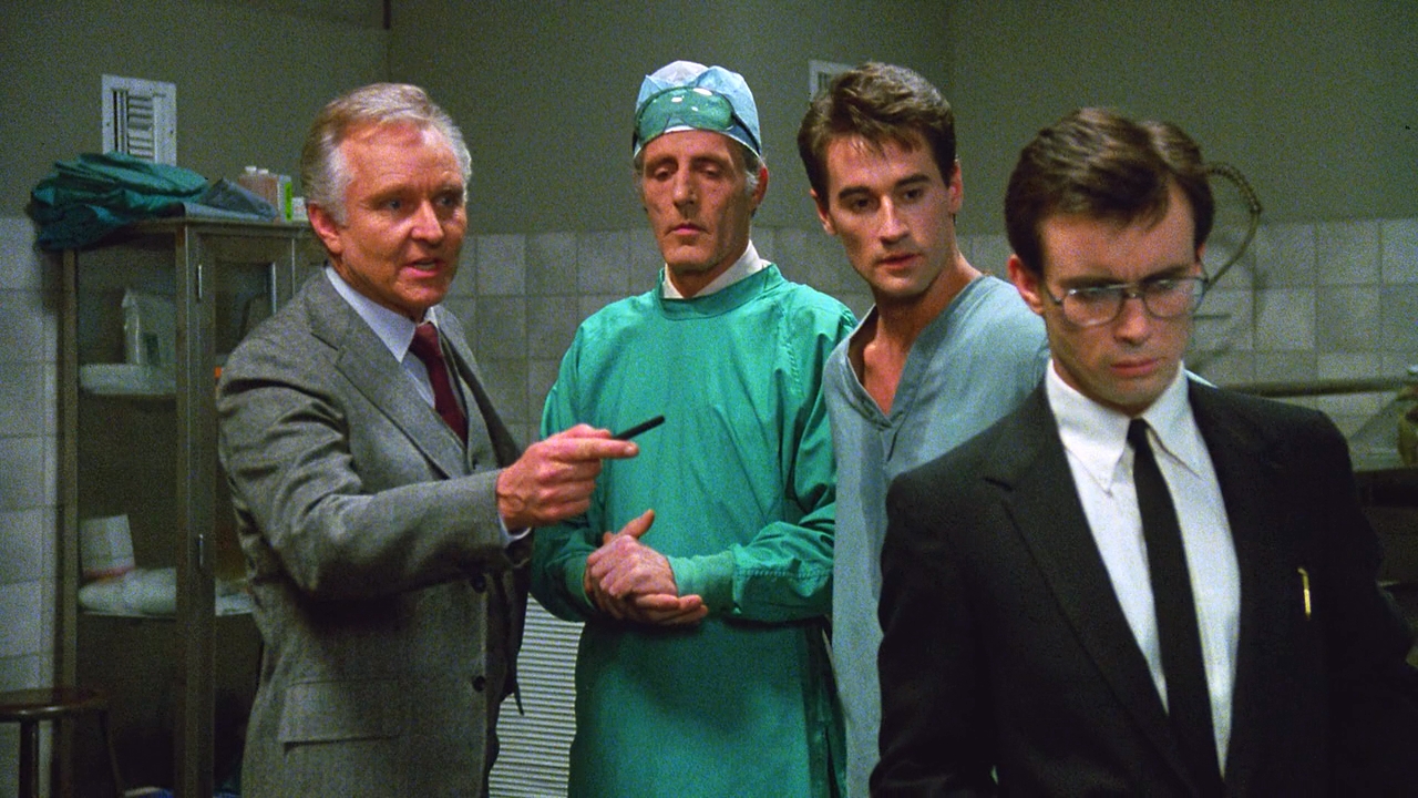 Four men stand in a lab gazing downwards at something just offscreen. Two are in suits, one in scrubs, and one looks like he just walked in from the street. One of the suits seems pissed.