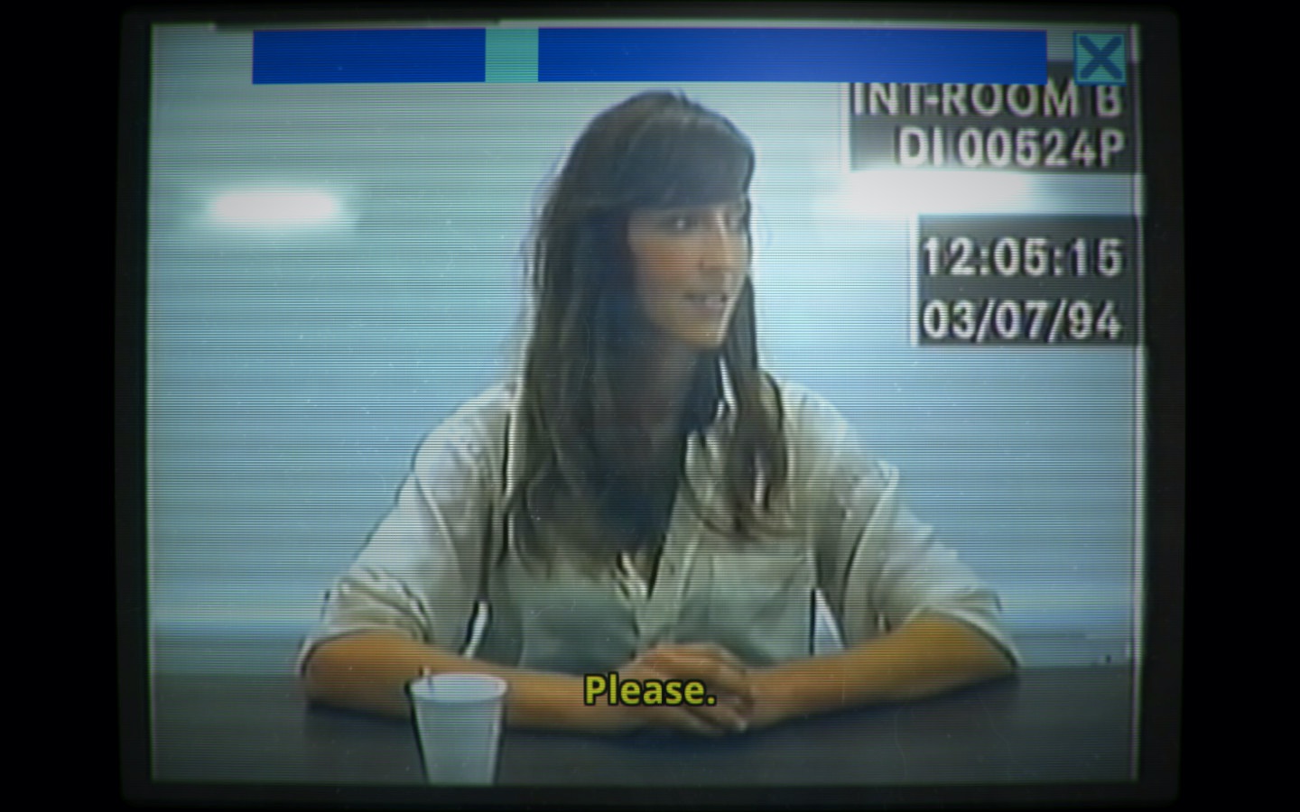 A still from the police interview videogame Her Story, presented as footage on an old CRT monitor. A young woman with long, dark hair sits at a table, looking slightly to the side with a faint smile.