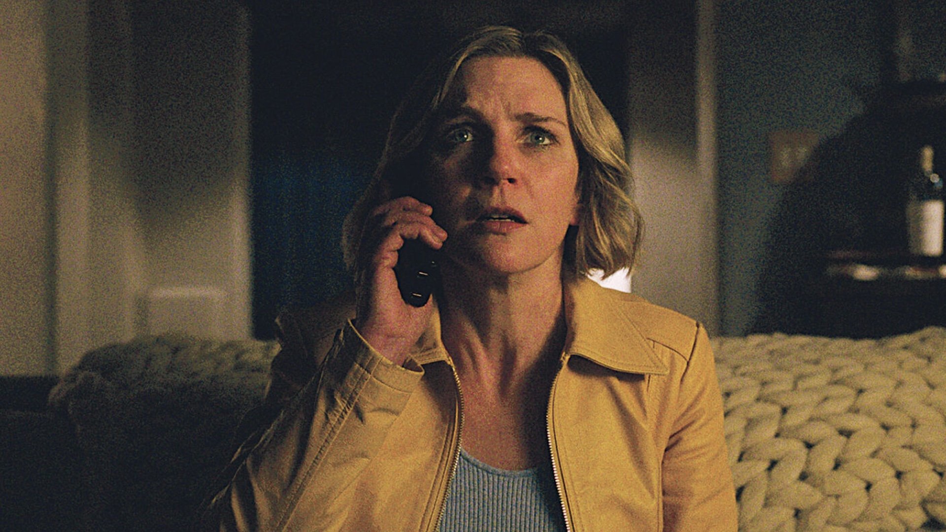 Carol, seated on the couch in her living room, holds a phone to her ear while staring straight ahead with a look of shock on her face.