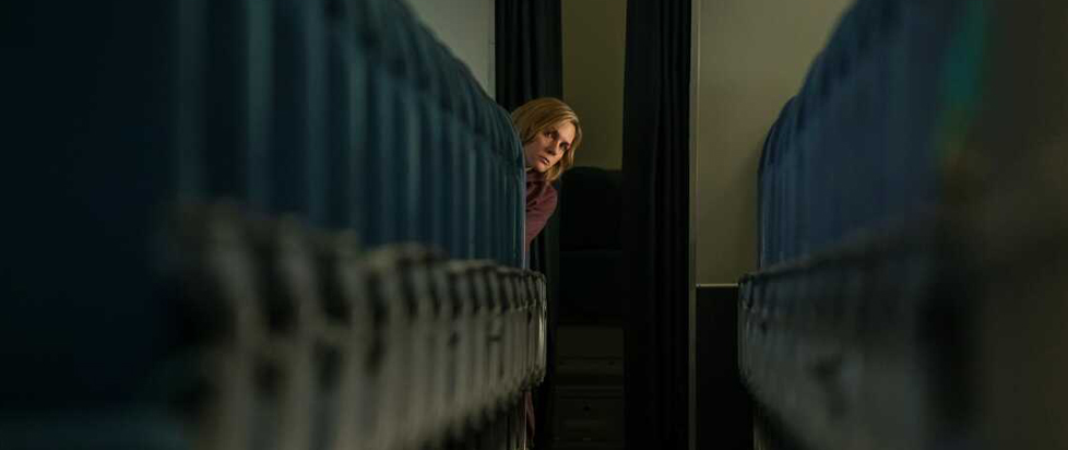 A low-angle, close-up shot down the aisle of an airplane shows actress Rhea Seehorn as the character Carol from the series Pluribus peeking her head out from behind one of the seats.