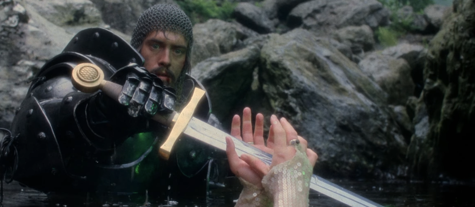 A screenshot from the trailer for Excalibur where Arthur in full armor is grasping the legendary sword from the hands of the Lady in the Lake