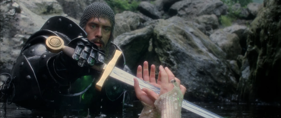 A screenshot from the trailer for Excalibur where Arthur in full armor is grasping the legendary sword from the hands of the Lady in the Lake