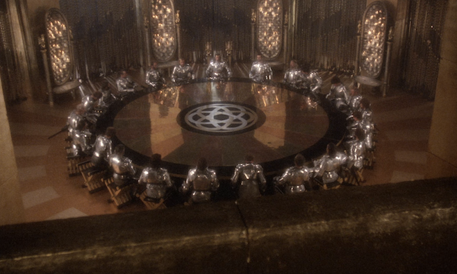 A screenshot from the trailer of Excalibur where Arthur has assembled his knights to the round table all sparkling in their silver armor and the room richly lacquered