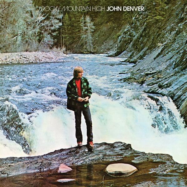 The cover of Rocky Mountain High by John Denver where the singer is in the center on a rock in front of a river with a short waterfall and trees and everything is lush and blue and green