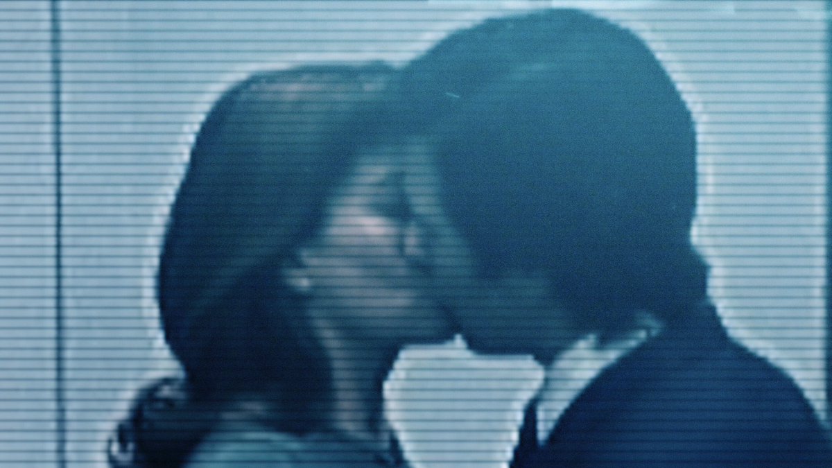 As seen on a closed-circuit monitor, Mark and Helly share a kiss.