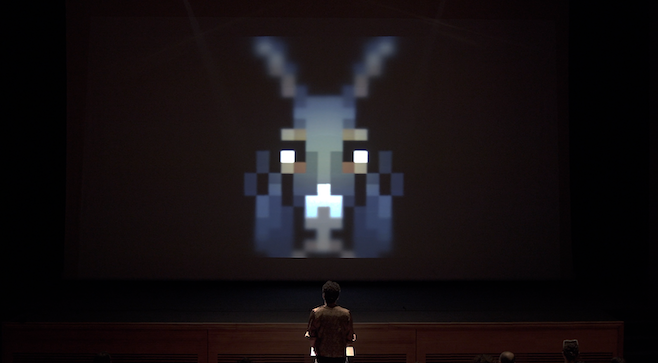 A photograph from asses.masses where the person at the podium is looking up at a projection of a large 8-bit donkey face