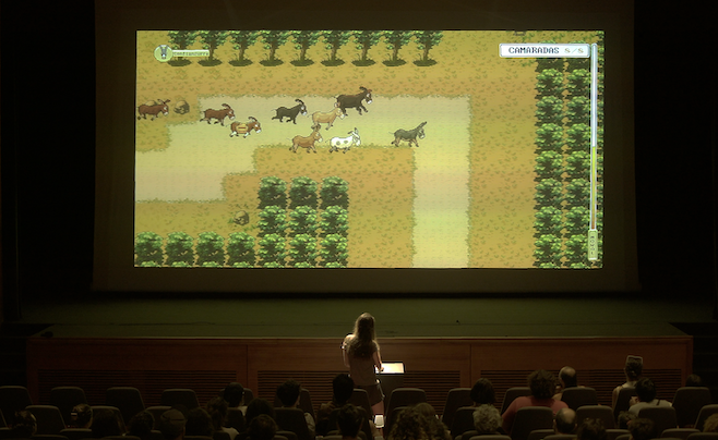 A photograph from asses masses where a main player at the front of an audience stands at a podium looking at a projection of a videogame of donkeys running around a little path
