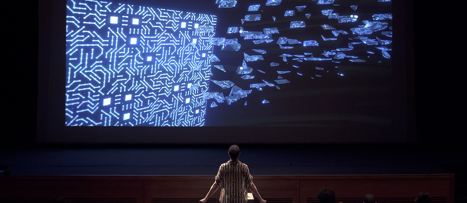 A photograph from asses.masses where someone is at the front of an audience watching blue lines on a projected screen