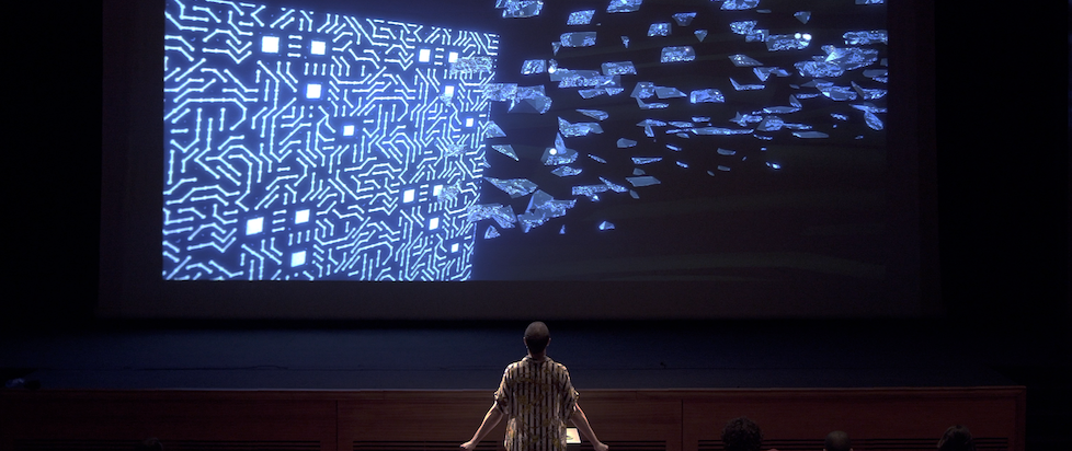 A photograph from asses.masses where someone is at the front of an audience watching blue lines on a projected screen