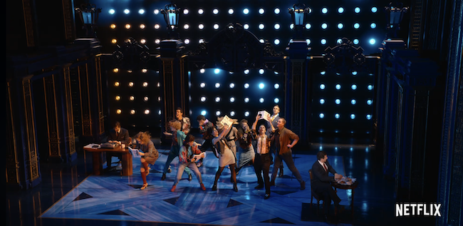 A screenshot from the Netflix trailer for Diana with a bunch of people on stage reading the news while a man in a suit tries to eat breakfast