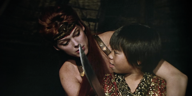 A screenshot from the trailer for Red Sonja where the barbarian woman is teaching little Eddie Reyes Jr. how to hold a sword but he's probably got it sorted already
