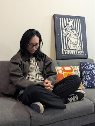 A photograph of Sam Eng with long black hair and round glasses sitting cross-legged on a couch with a print of a rabbit on a skateboard behind 