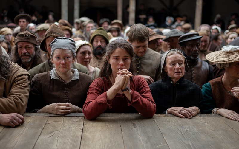 Agnes leans on the edge of the stage at The Globe, hands clasped in prayer.