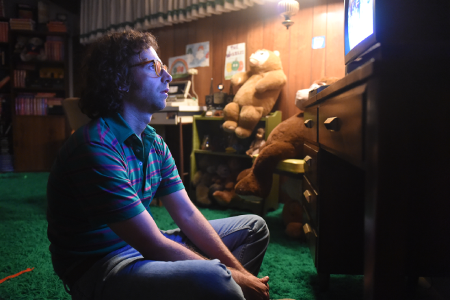 A screenshot from Bigsby Bear where a young person is sitting criss cross applesauce on green carpet staring at a CRT tv and surrounded by childish drawings and stuffed bears