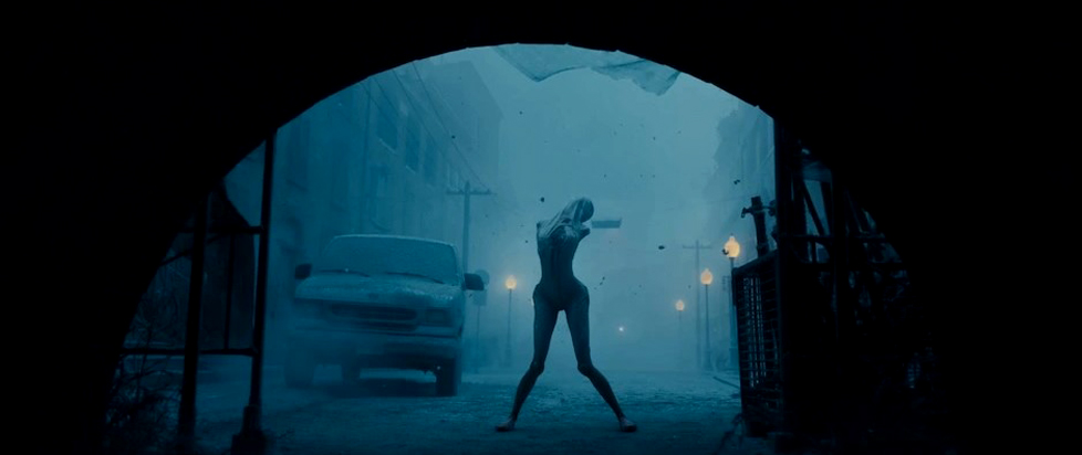 A still from Return to Silent Hill shows a woman with her arms and legs bent at unnatural angles stands beneath the arch of an overpass on a gloomy, foggy day.
