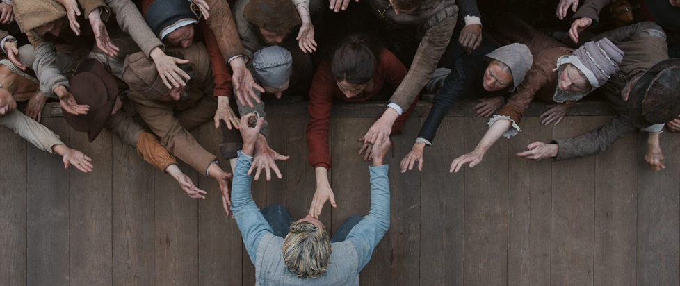 A still from Hamnet shows a top-down view of an actor onstage holding his hands out towards the audience as they reach towards him in turn.
