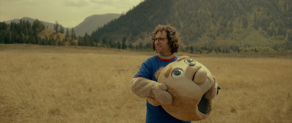 A screenshot from Bigsby Bear where a young person in a bear mascot costume with a blue shirt on is holding the bear head mask and standing in yellow grass by some mountains