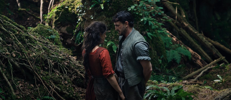 An image from Hamnet where William in rolled up sleeves on the right and his wife in red on the left are standing in the woods looking at each other