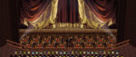 A screenshot from Final Fantasy VI shows large, ornate red and gold curtains hanging prominently above a multi-tiered orchestra pit. Two levels of musicians are seen playing instruments, led by a conductor positioned in the center.