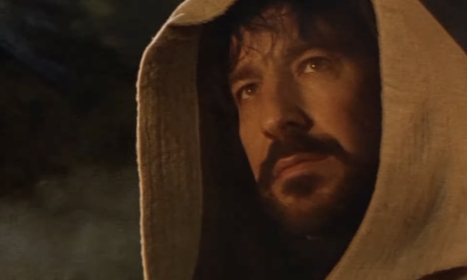 A screenshot from the trailer of Robin Hood Prince of Thieves where Alan Rickman with dark hair and beard under a white clandestine rob stares with firelight playing across his face